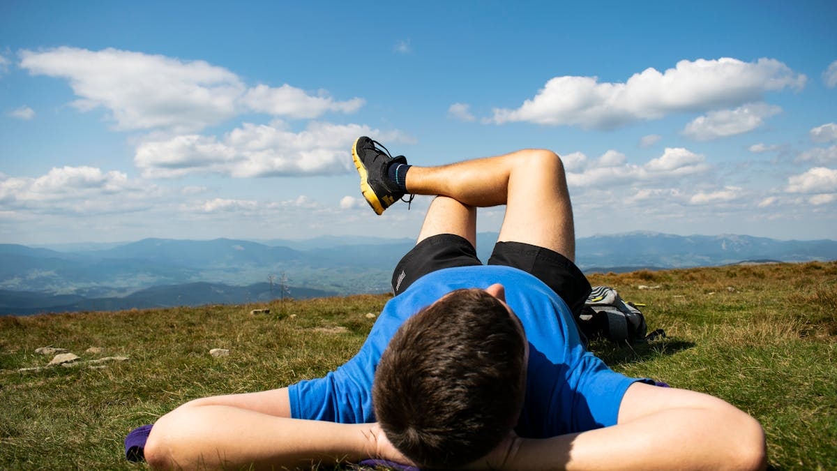 Unrecognizable young male athlete in sportswear lying on grassy ground in mountainous terrain and resting after outdoor workout
